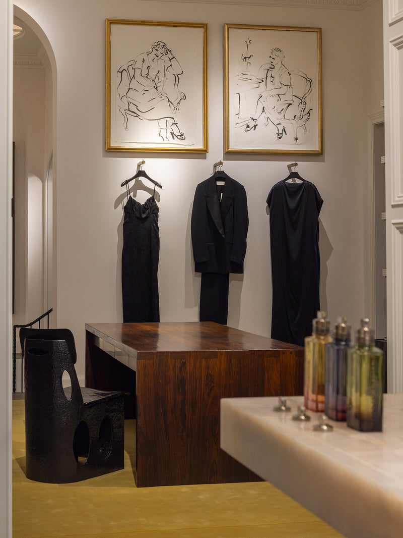 A styled interior room with three black garments hanging on hangers against a cream wall beneath two framed line drawings. A dark wooden table and chair are in the foreground, with fragrance bottles on a counter in the lower right.