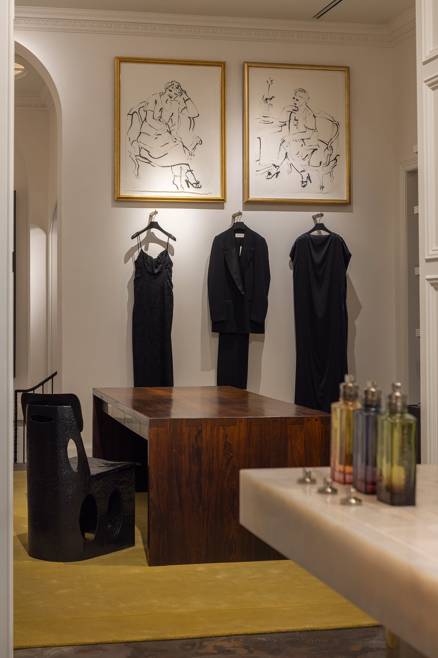 A styled interior room with three black garments hanging on hangers against a cream wall beneath two framed line drawings. A dark wooden table and chair are in the foreground, with fragrance bottles on a counter in the lower right.