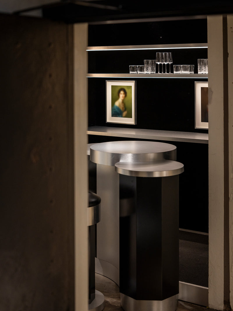 A modern cylindrical bar table with a black body and brushed silver metallic top and base, positioned in front of a dark shelving unit with glasses and framed art.