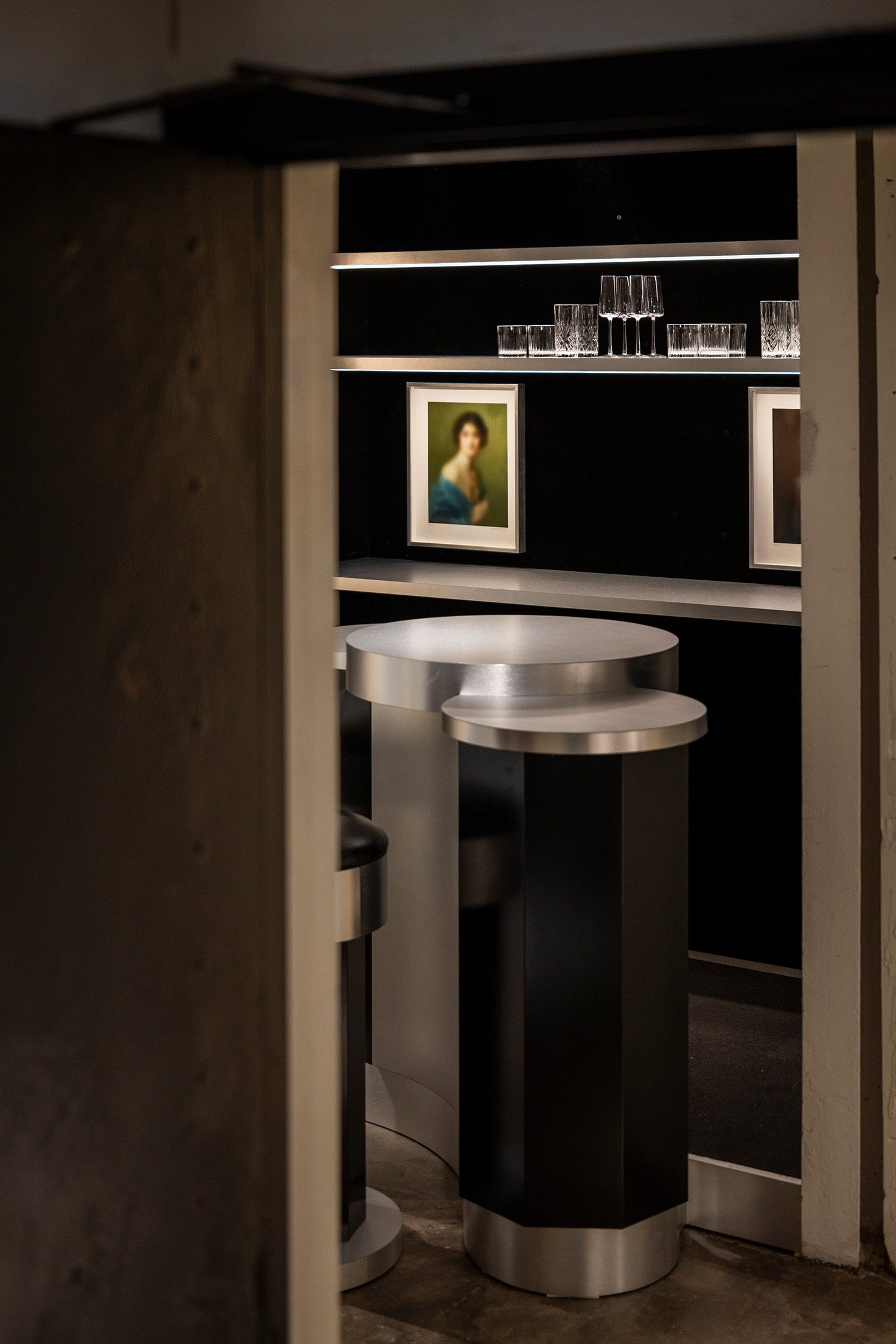 A modern cylindrical bar table with a black body and brushed silver metallic top and base, positioned in front of a dark shelving unit with glasses and framed art.