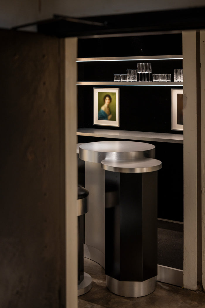 A modern cylindrical bar table with a black body and brushed silver metallic top and base, positioned in front of a dark shelving unit with glasses and framed art.