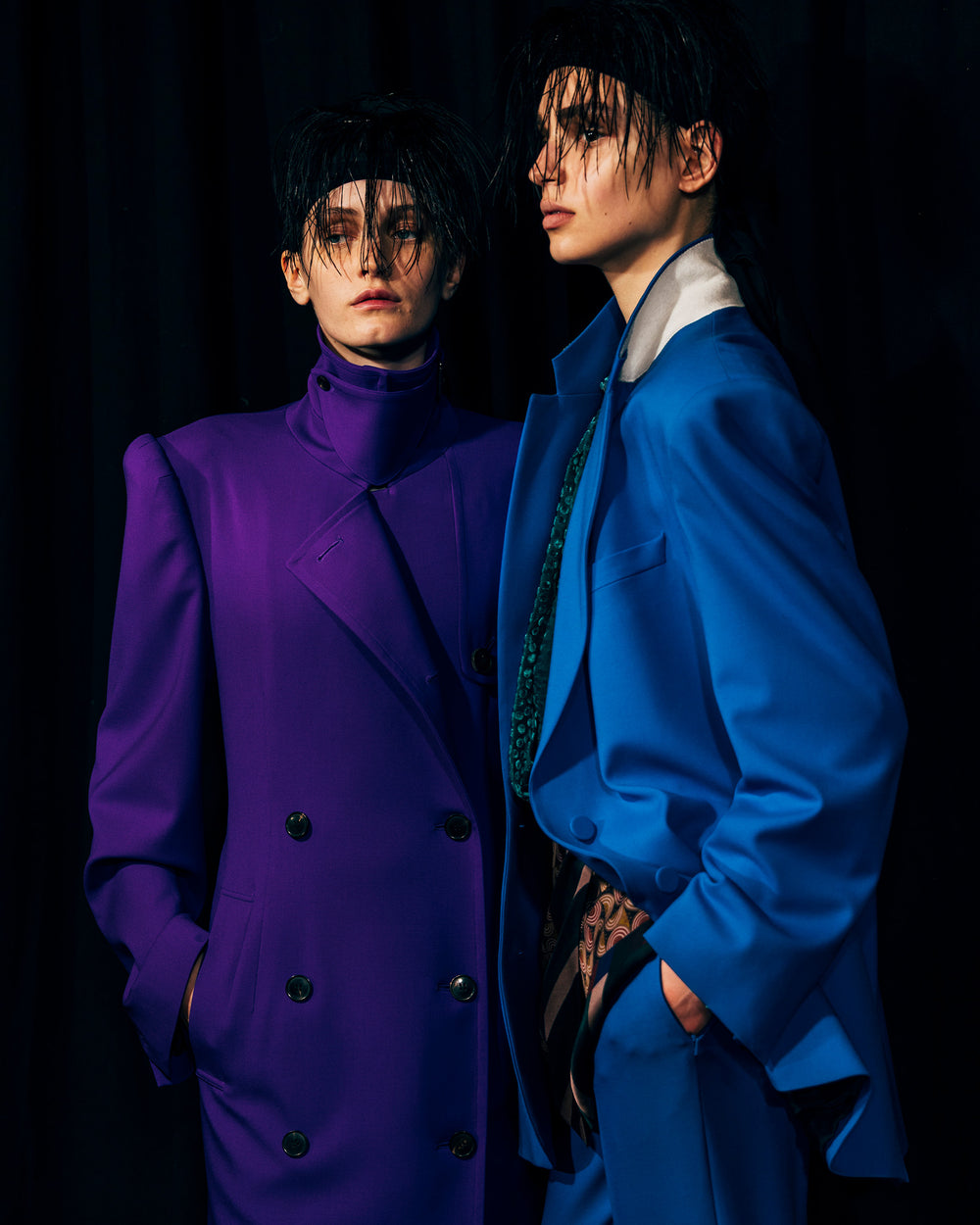 Two individuals wearing vibrant purple and blue suits against a black background