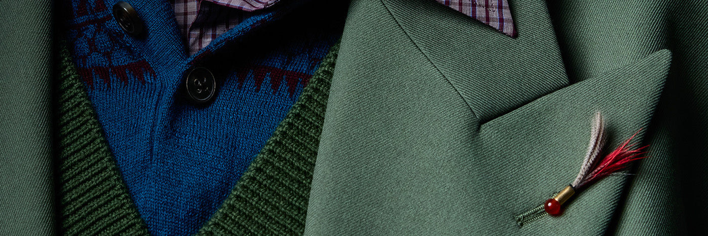 Close-up of a tailored olive green suit jacket with a feathered lapel pin, layered over a blue knit sweater vest and a patterned collared shirt.