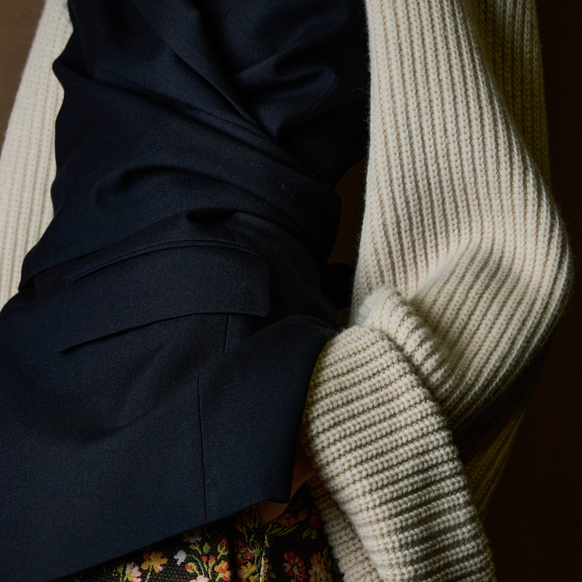 Close-up of a person wearing a dark navy blazer with a cream ribbed knit sleeves, with a floral garment visible at the bottom.