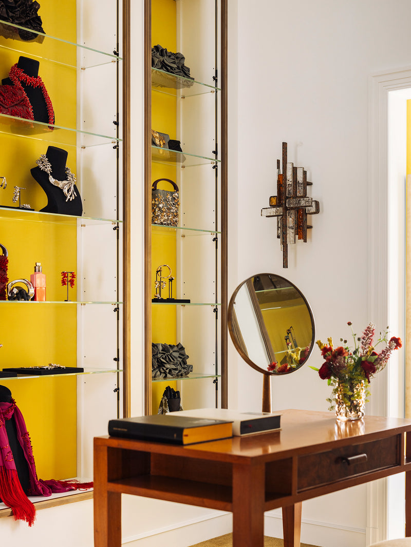 A styled interior room with a wooden vanity desk with a round mirror, books, and a vase of flowers and in the back tall glass display cabinets set against bright yellow wall, showcasing jewelry and accessories. 