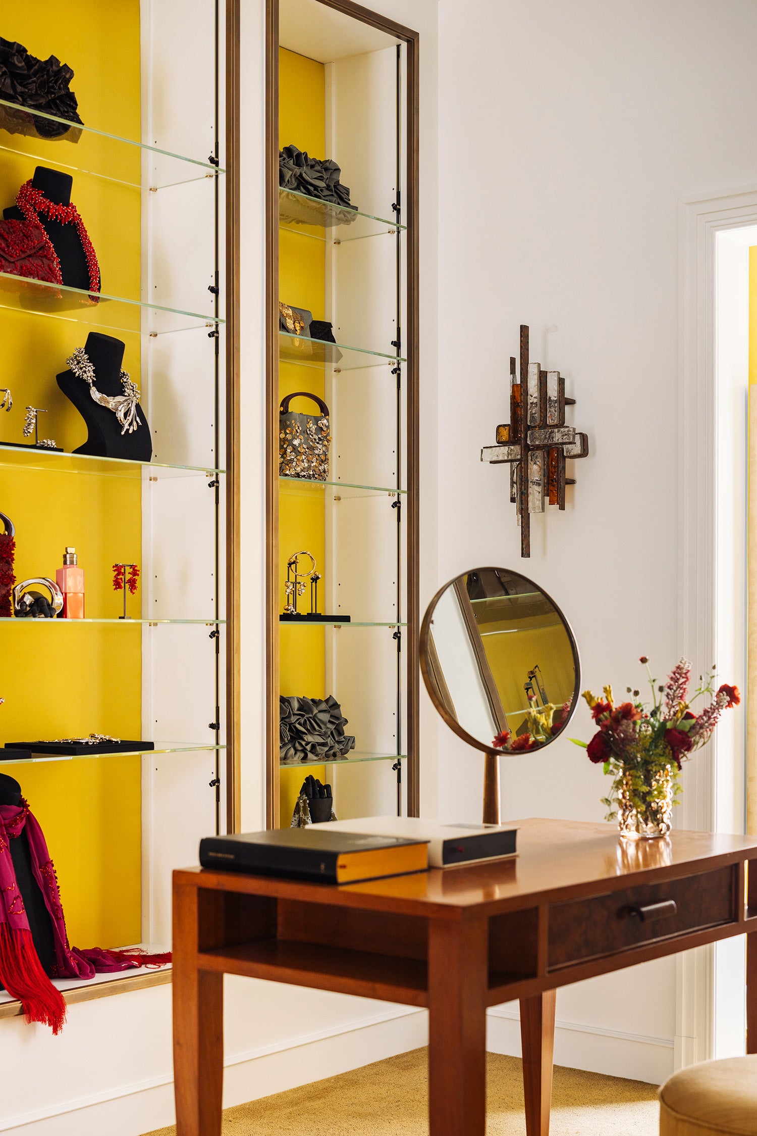 A styled interior room with a wooden vanity desk with a round mirror, books, and a vase of flowers and in the back tall glass display cabinets set against bright yellow wall, showcasing jewelry and accessories. 