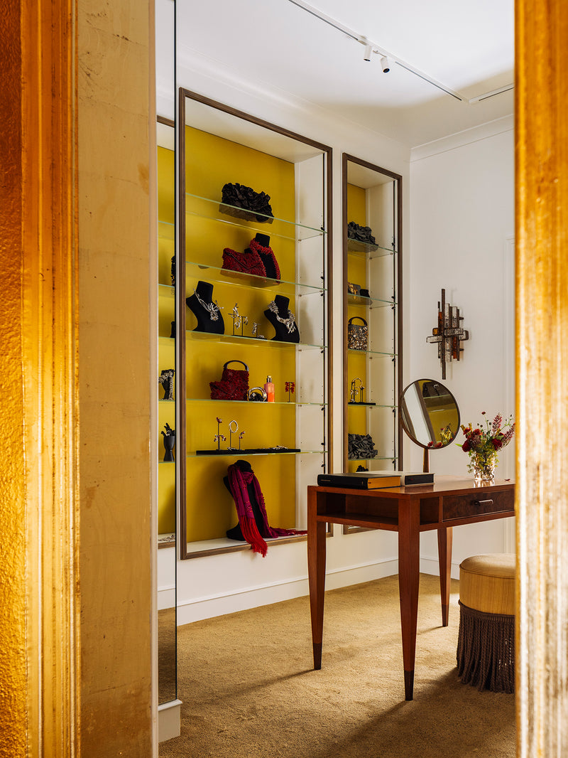 A luxury interior room featuring a vanity desk with an oval mirror, a yellow accent wall with glass display shelves, and a stool, framed by a textured gold column in the foreground.