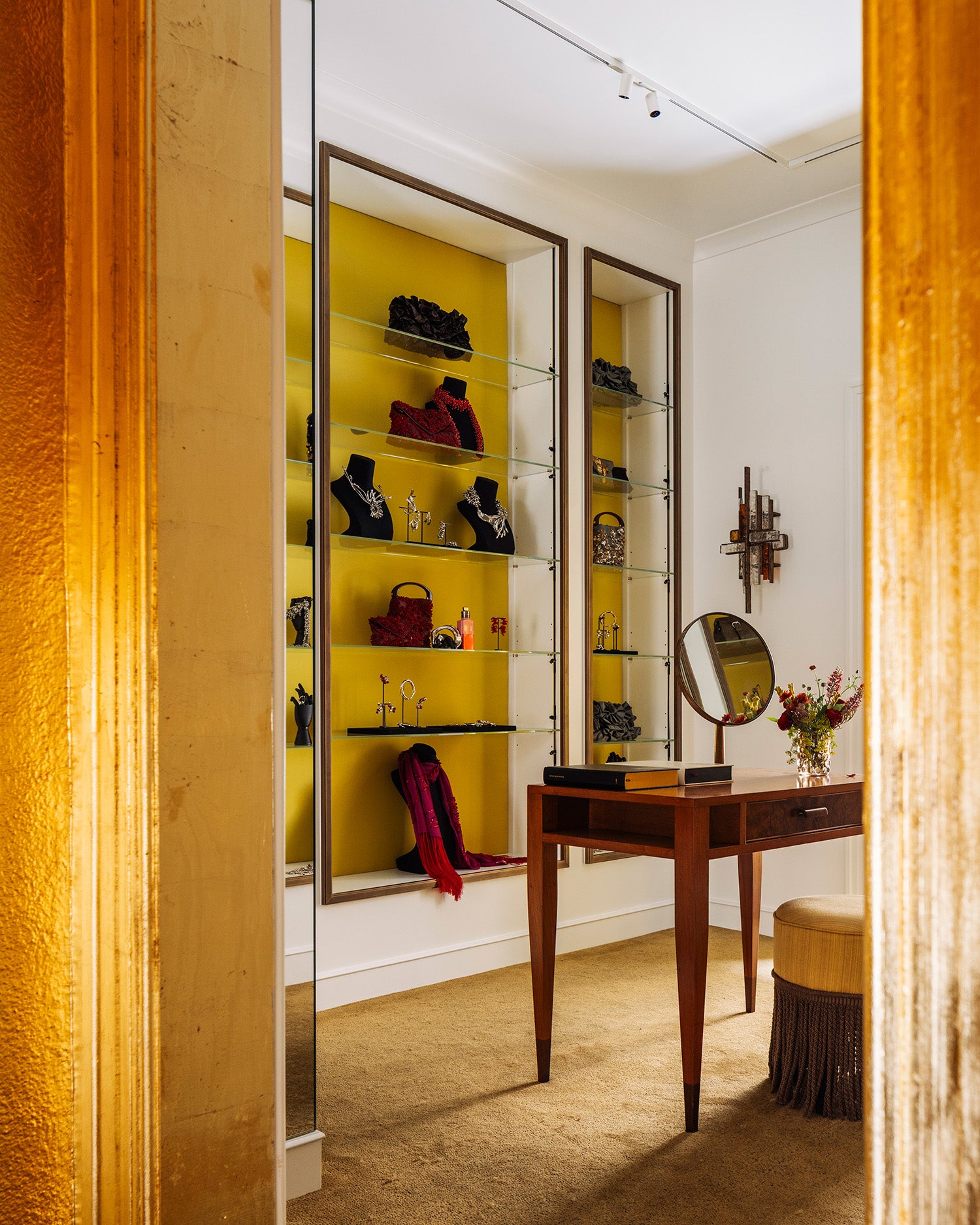 A luxury interior room featuring a vanity desk with an oval mirror, a yellow accent wall with glass display shelves, and a stool, framed by a textured gold column in the foreground.