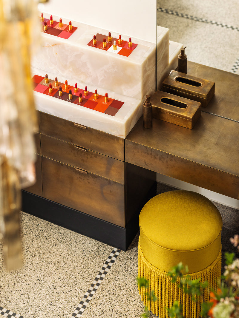 A styled interior scene showing a bronze vanity having a white marble area with lipstick bullets, one bronze tissue box, and a round mustard-yellow fringed stool on a speckled floor.