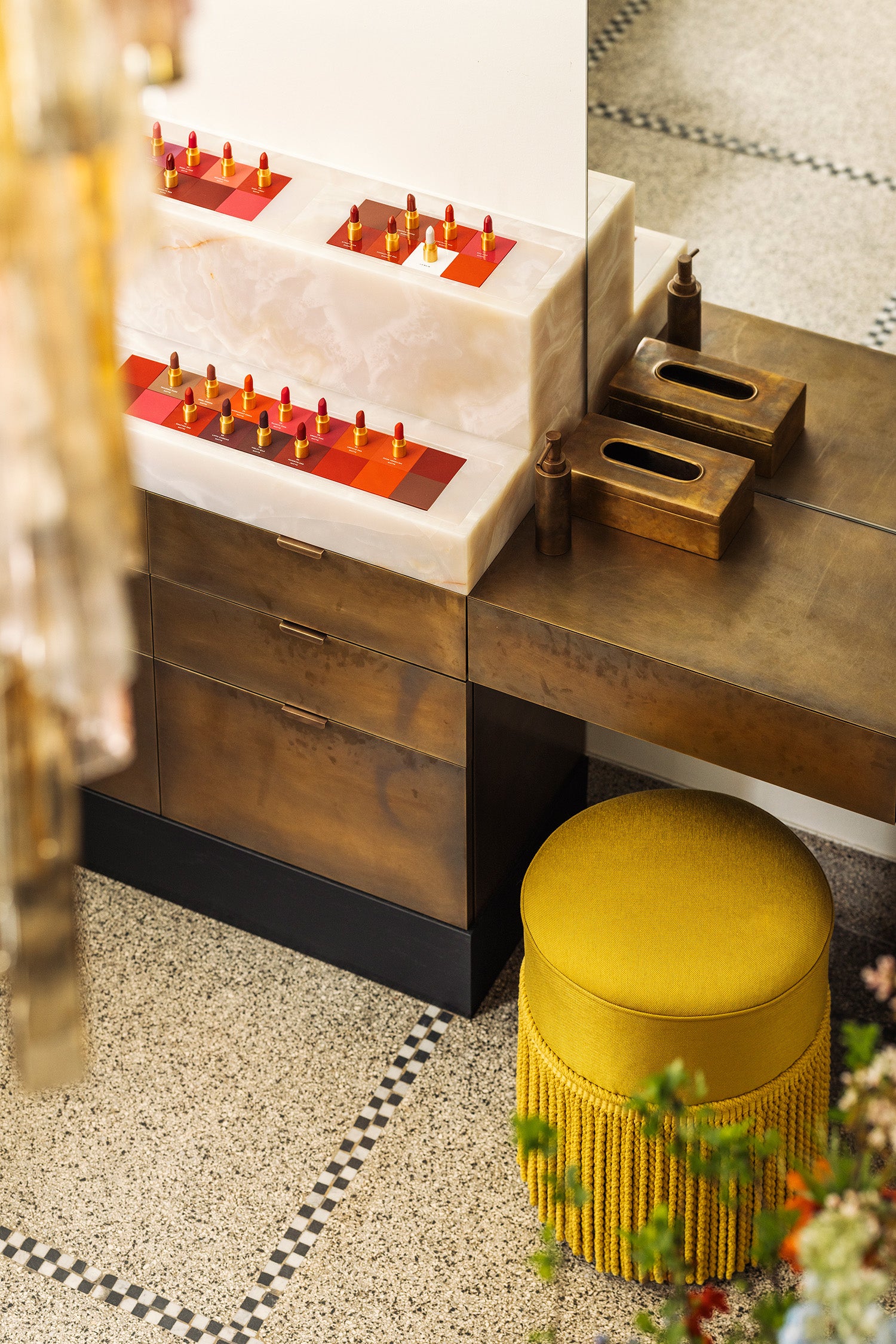 A styled interior scene showing a bronze vanity having a white marble area with lipstick bullets, one bronze tissue box, and a round mustard-yellow fringed stool on a speckled floor.