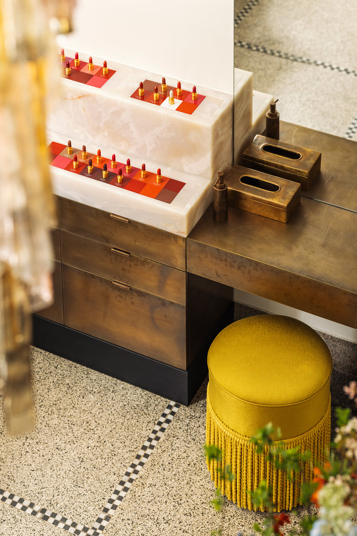 A styled interior scene showing a bronze vanity having a white marble area with lipstick bullets, one bronze tissue box, and a round mustard-yellow fringed stool on a speckled floor.