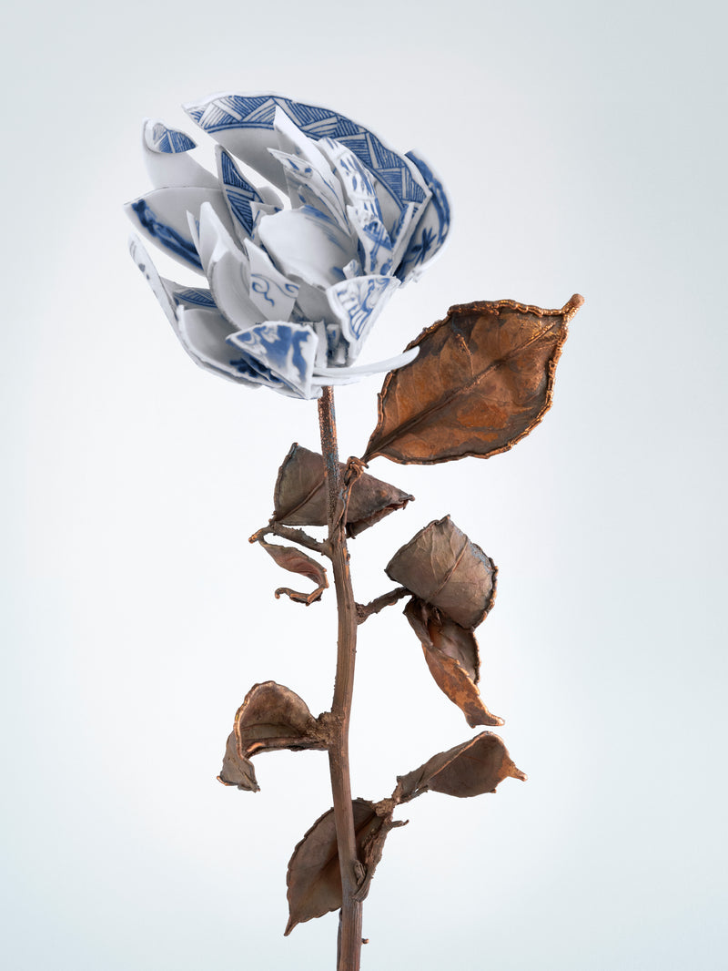 White ceramic flower with blue patterns on a stem with dried leaves against a light gray background - Dries Van Noten