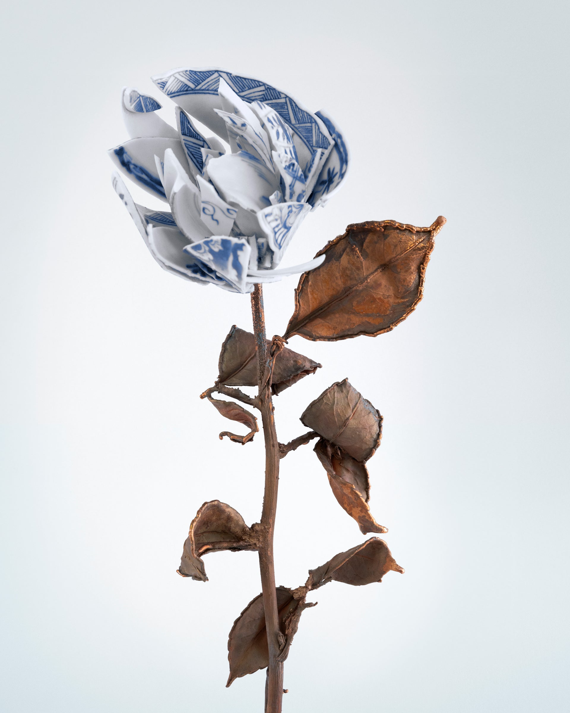 White ceramic flower with blue patterns on a stem with dried leaves against a light gray background - Dries Van Noten