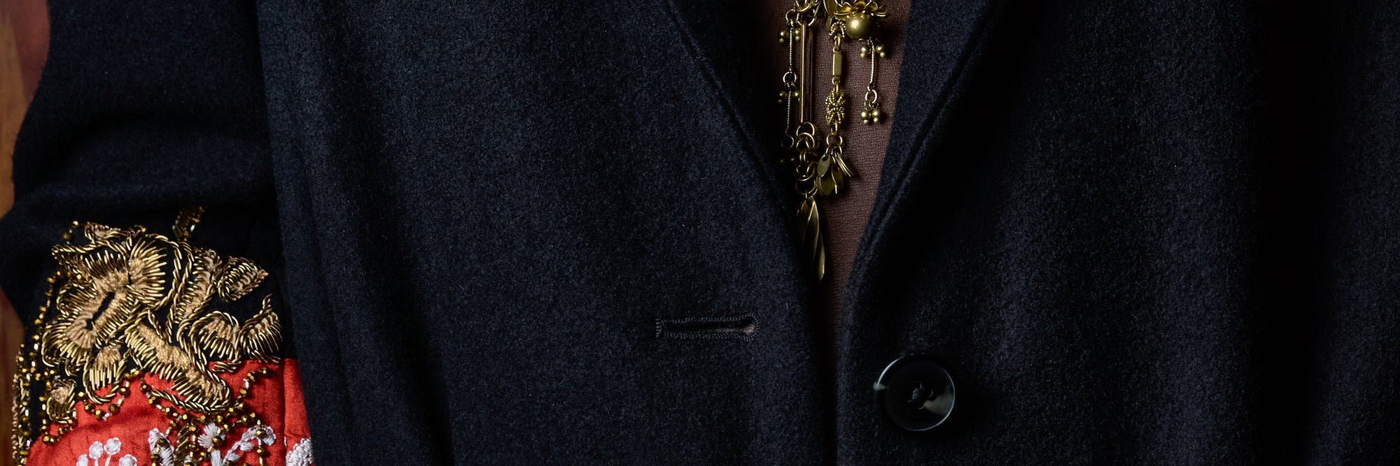 Close-up of a dark navy wool-blend blazer with a single button closure, featuring a gold chain necklace and a red and gold embroidered sleeve detail.