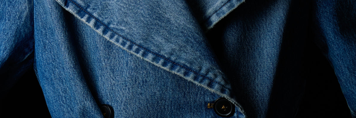 Close-up of a blue denim jacket with visible fabric texture, a pointed lapel, and dark buttons against a dark background.