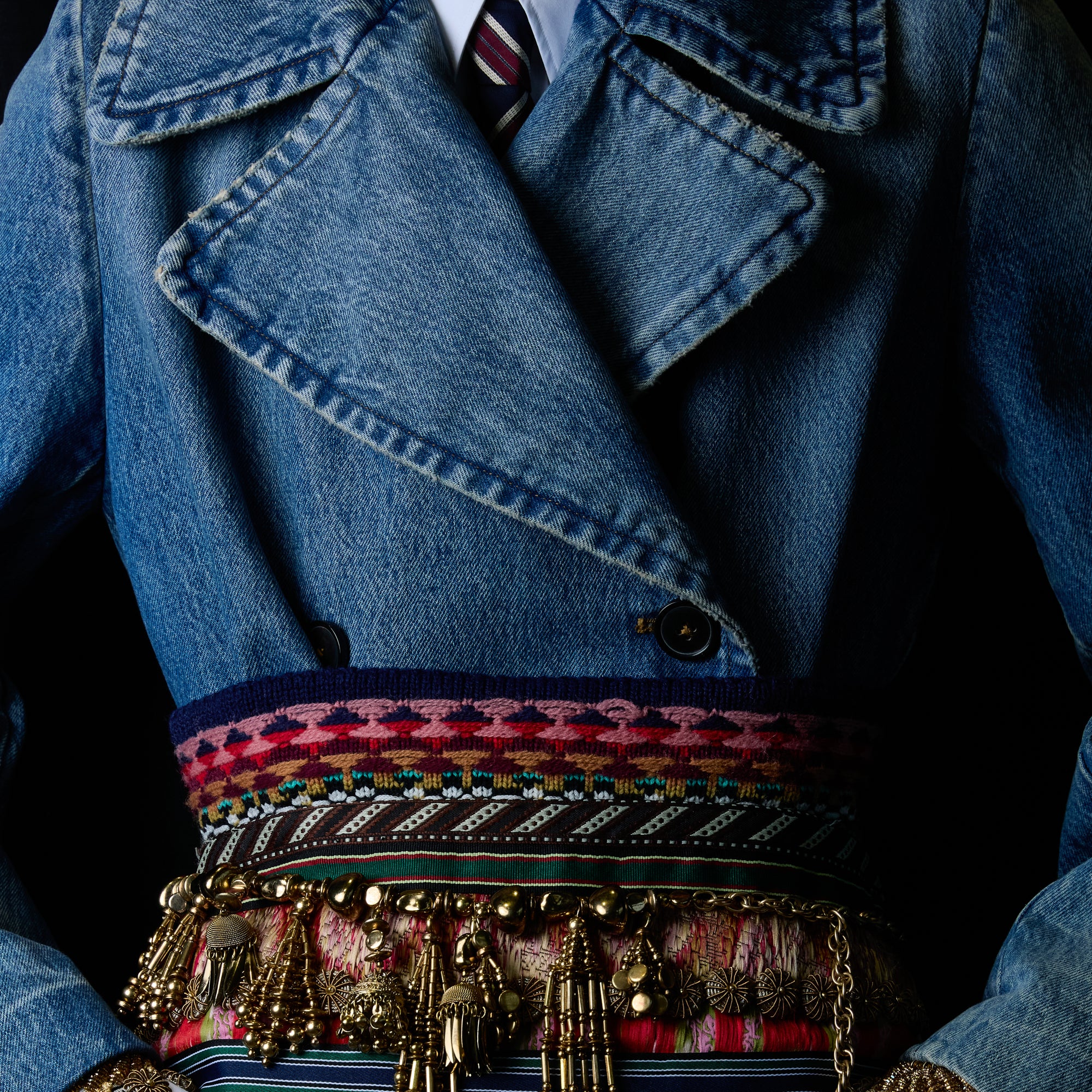 A close-up of a person wearing a blue denim jacket with a patterned multicolor waist belt with metallic coin charms, a striped tie, against a dark background.