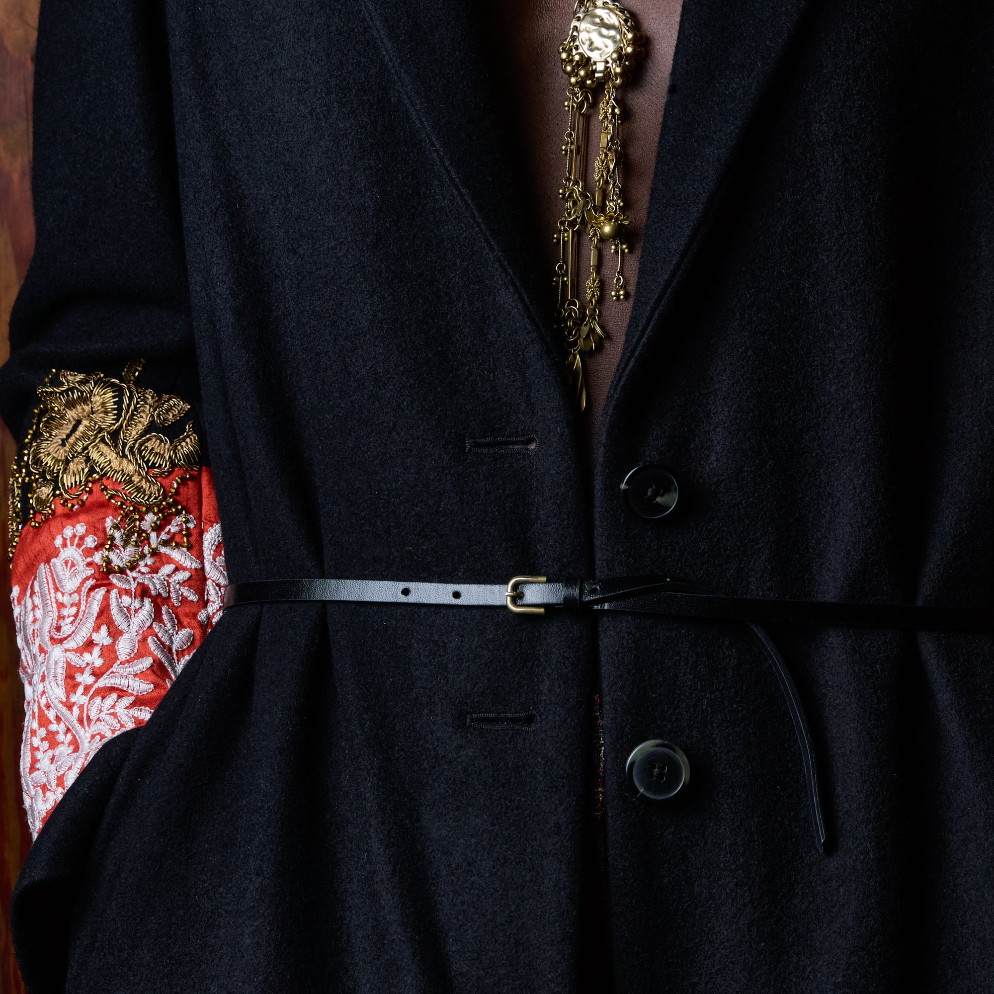 Close-up of a person wearing a dark navy wool-blend blazer with a thin black belt at the waist, a gold chain necklace, and a red-and-white patterned sleeve detail.