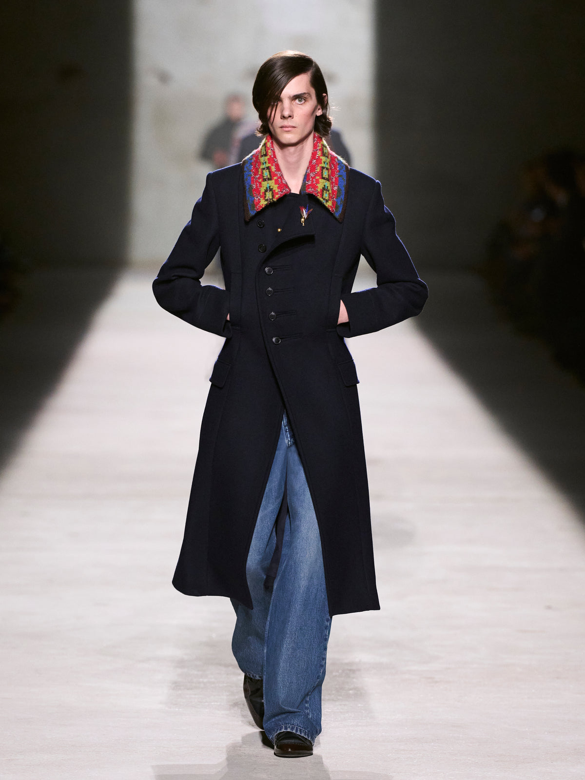 Model walking on a runway wearing a long dark coat with a colorful colar - Dries Van Noten