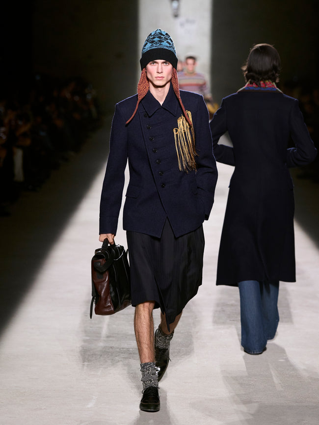 Model walking on a runway wearing a navy coat and hat with braided hair, carrying a leather bag - Dries Van Noten
