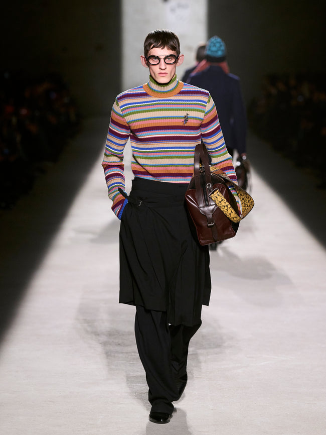 Model walking on a runway wearing a colorful striped sweater and black pants, carrying a brown leather bag - Dries Van Noten