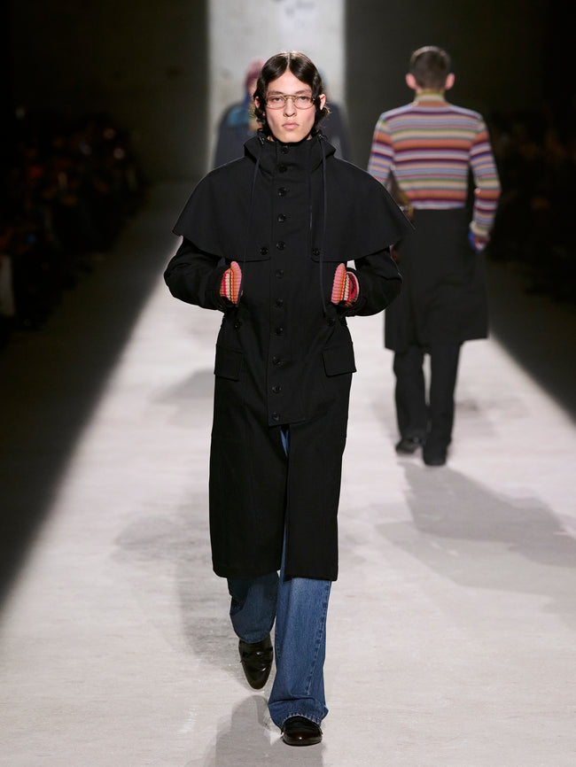 Model walking on a runway wearing a black coat and blue jeans - Dries Van Noten