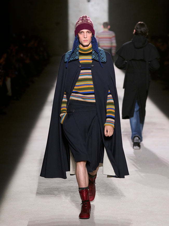 Model walking on a runway wearing a long dark coat, colorful striped sweater, and red boots - Dries Van Noten