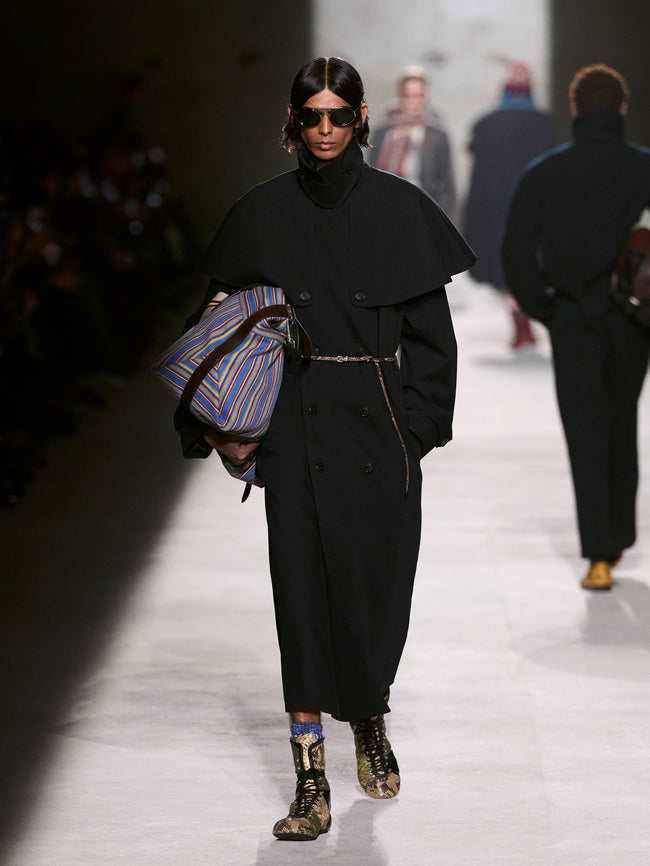 Model walking on a runway wearing a black outfit with a striped bag - Dries Van Noten