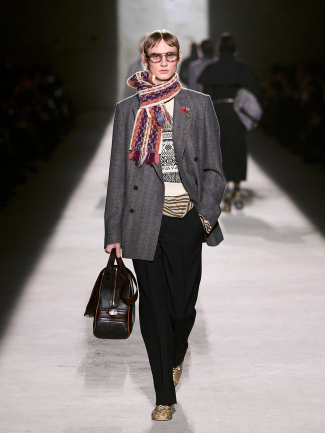 Model walking on a runway wearing a gray coat, patterned scarf, and holding a brown bag - Dries Van Noten