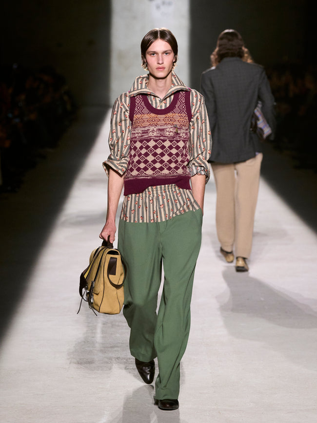 Model walking on a runway wearing a patterned sweater and green pants, holding a beige bag - Dries Van Noten