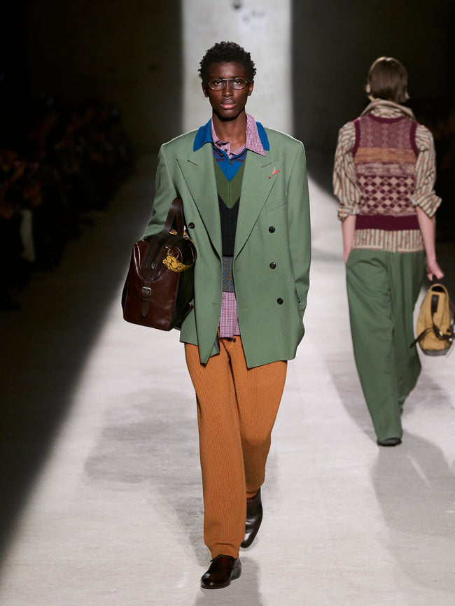 Two models walking on a runway, one in a green coat and brown pants, the other in a patterned top and green pants - Dries Van Noten
