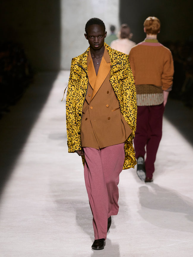 Model walking on a runway wearing a yellow floral coat over a brown suit with pink pants - Dries Van Noten