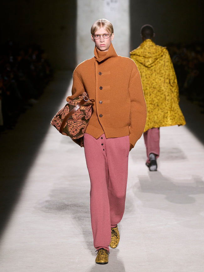 Model walking on a runway wearing a brown jacket and pink pants with a patterned bag - Dries Van Noten