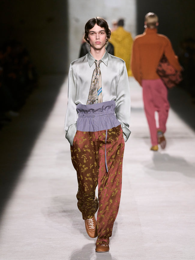 Model walking on a runway wearing a light gray shirt, patterned tie, and brown pants - Dries Van Noten
