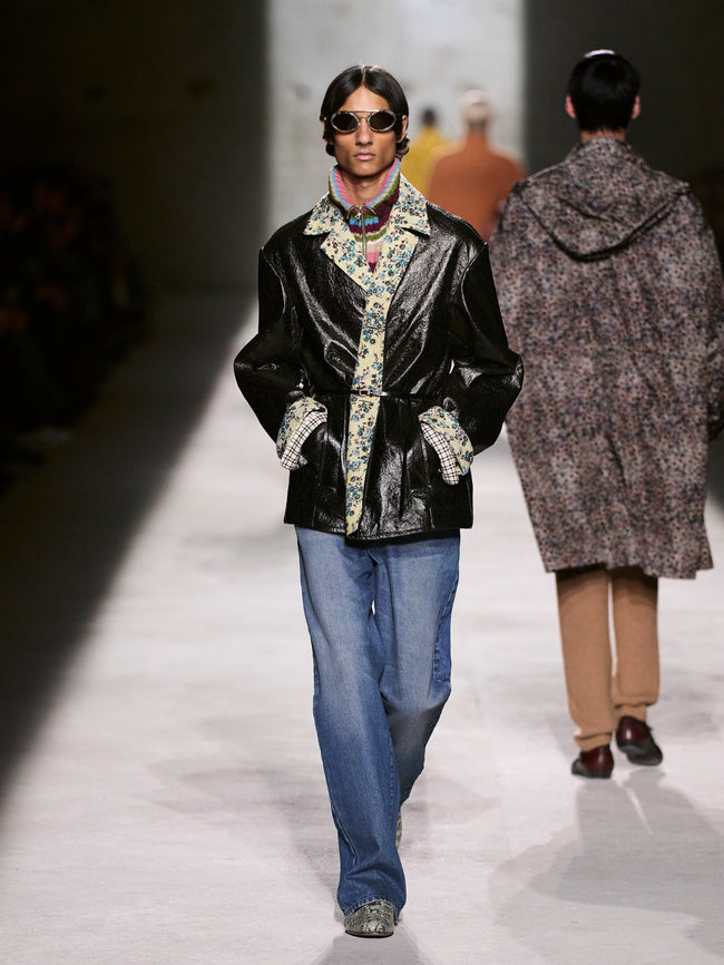 Model walking on a runway wearing a black jacket with floral patterns, blue jeans, and sunglasses - Dries Van Noten