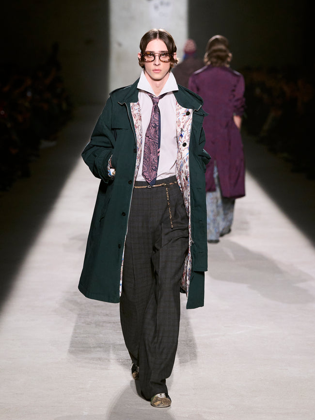 Model walking on a runway wearing a long dark coat and patterned tie - Dries Van Noten