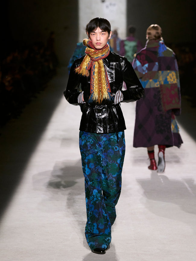 Model walking on a runway wearing a black jacket, colorful scarf, and blue floral pants - Dries Van Noten