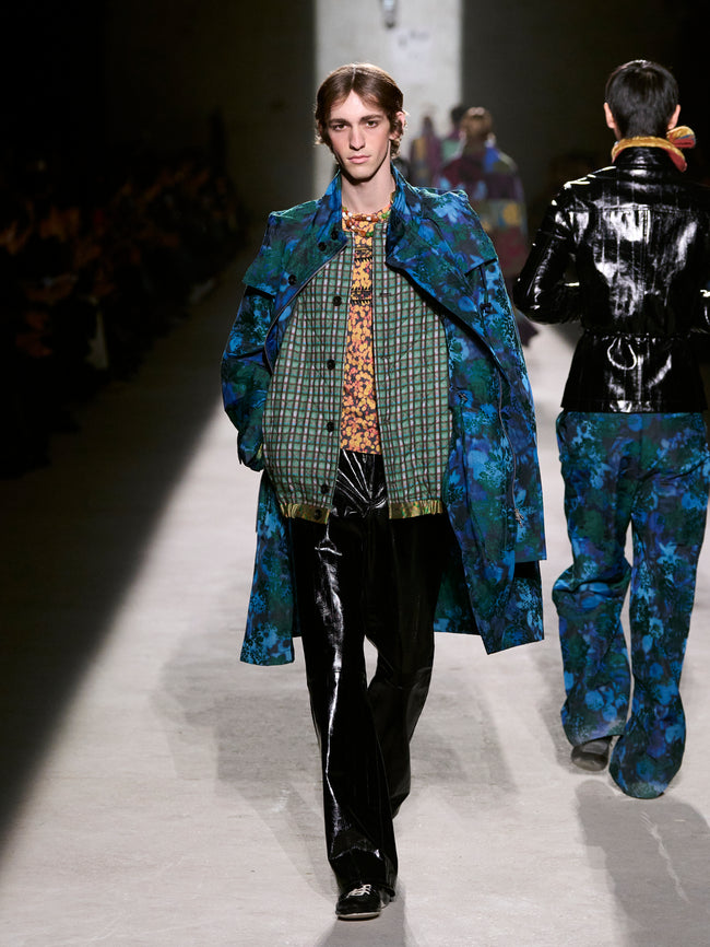Model walking on a runway wearing a blue patterned coat and black pants- Dries Van Noten
