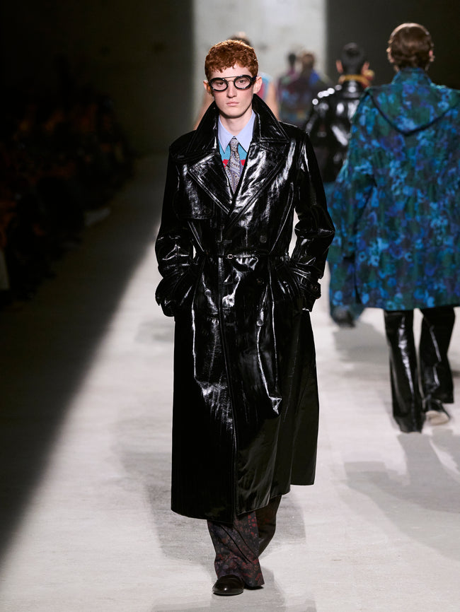 Model wearing a black glossy coat on a runway with another model in a blue floral coat in the background - Dries Van Noten