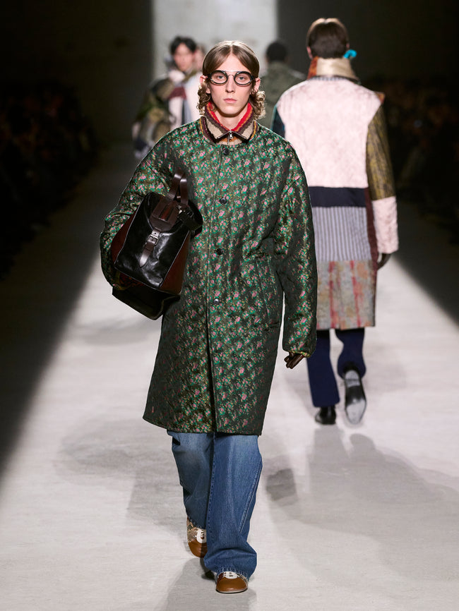 Model walking on a runway wearing a green patterned coat and blue jeans - Dries Van Noten