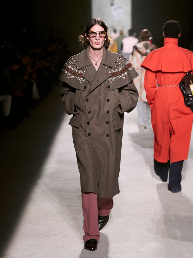 Model walking on a runway wearing a long coat with embellishments - Dries Van Noten