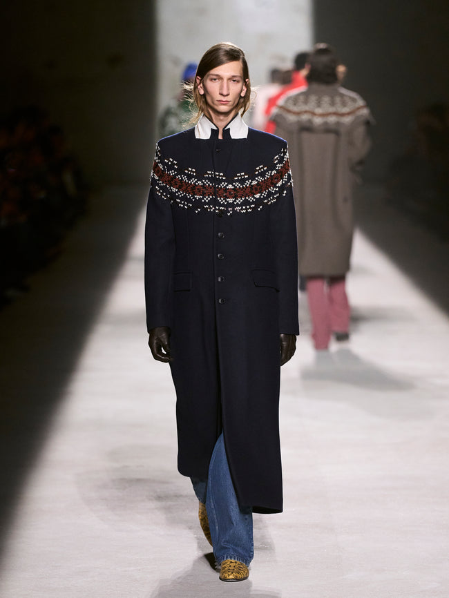 Model walking on a runway wearing a long dark coat with a patterned sweater - Dries Van Noten