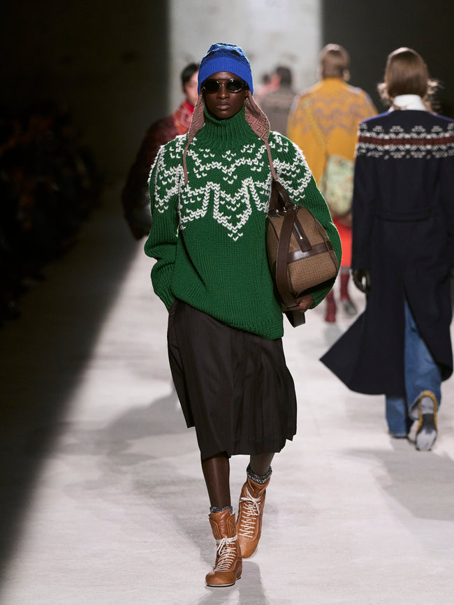 Model walking on a runway wearing a green sweater with white patterns, black skirt, and brown boots - Dries Van Noten