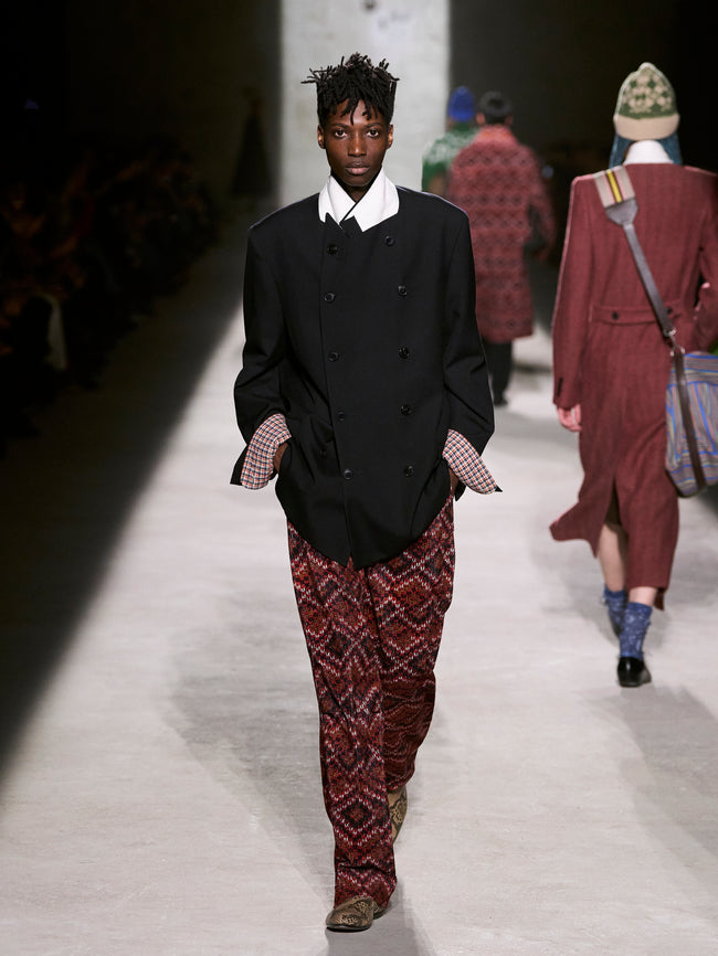 Model walking on a runway wearing a black blazer and patterned pants - Dries Van Noten