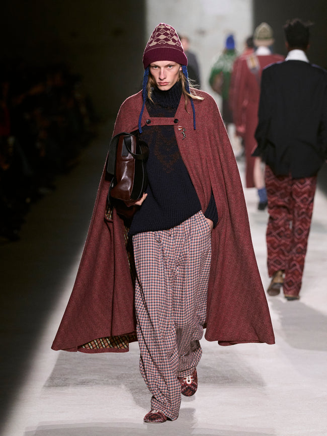 Model walking on a runway wearing a long burgundy coat, patterned hat, and plaid pants - Dries Van Noten