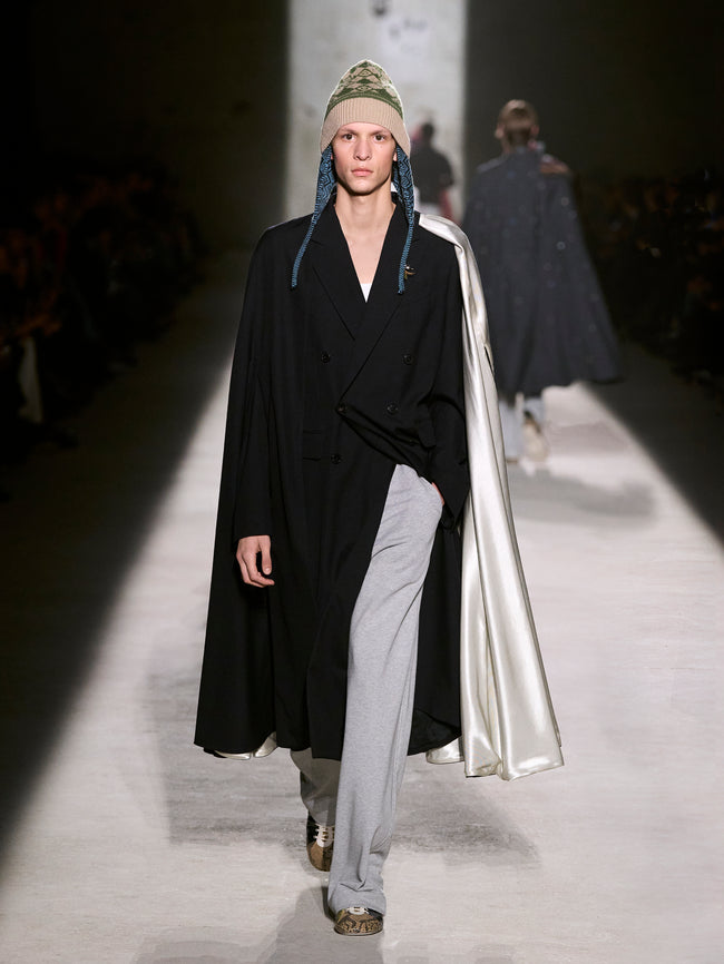 Model walking on a runway wearing a black coat with a silver lining and a green beanie - Dries Van Noten