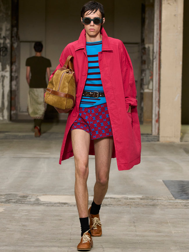 Person wearing a red coat, blue striped shirt, and patterned shorts walking outdoors. - Dries Van Noten