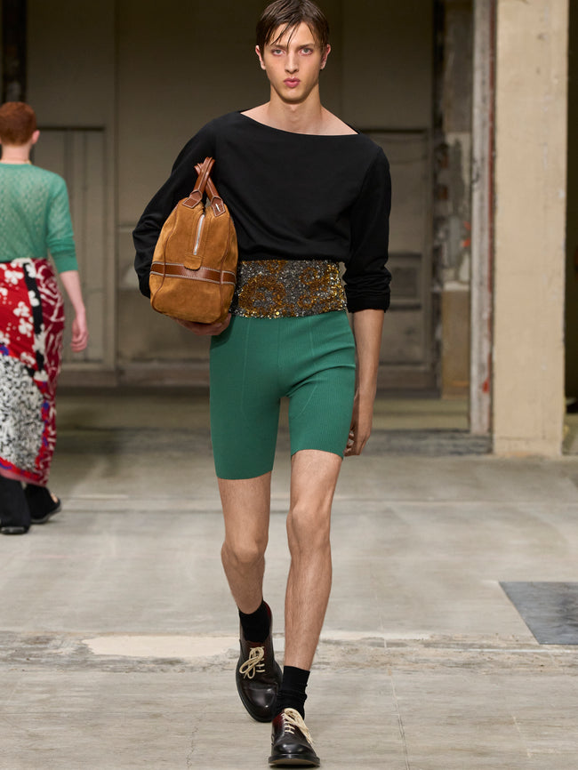 Model walking on a runway wearing a black top, green shorts, and carrying a brown bag. - Dries Van Noten
