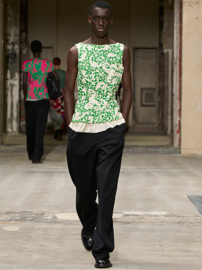 Model wearing a green floral sleeveless top and black pants walking outdoors. - Dries Van Noten