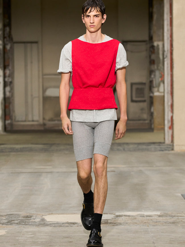 Model wearing a red vest over a white shirt with gray shorts on a runway. - Dries Van Noten