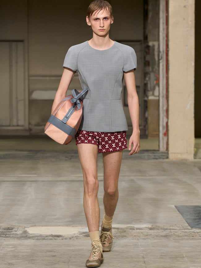 Man walking with a bag on a wooden floor, wearing a gray t-shirt and patterned shorts. - Dries Van Noten